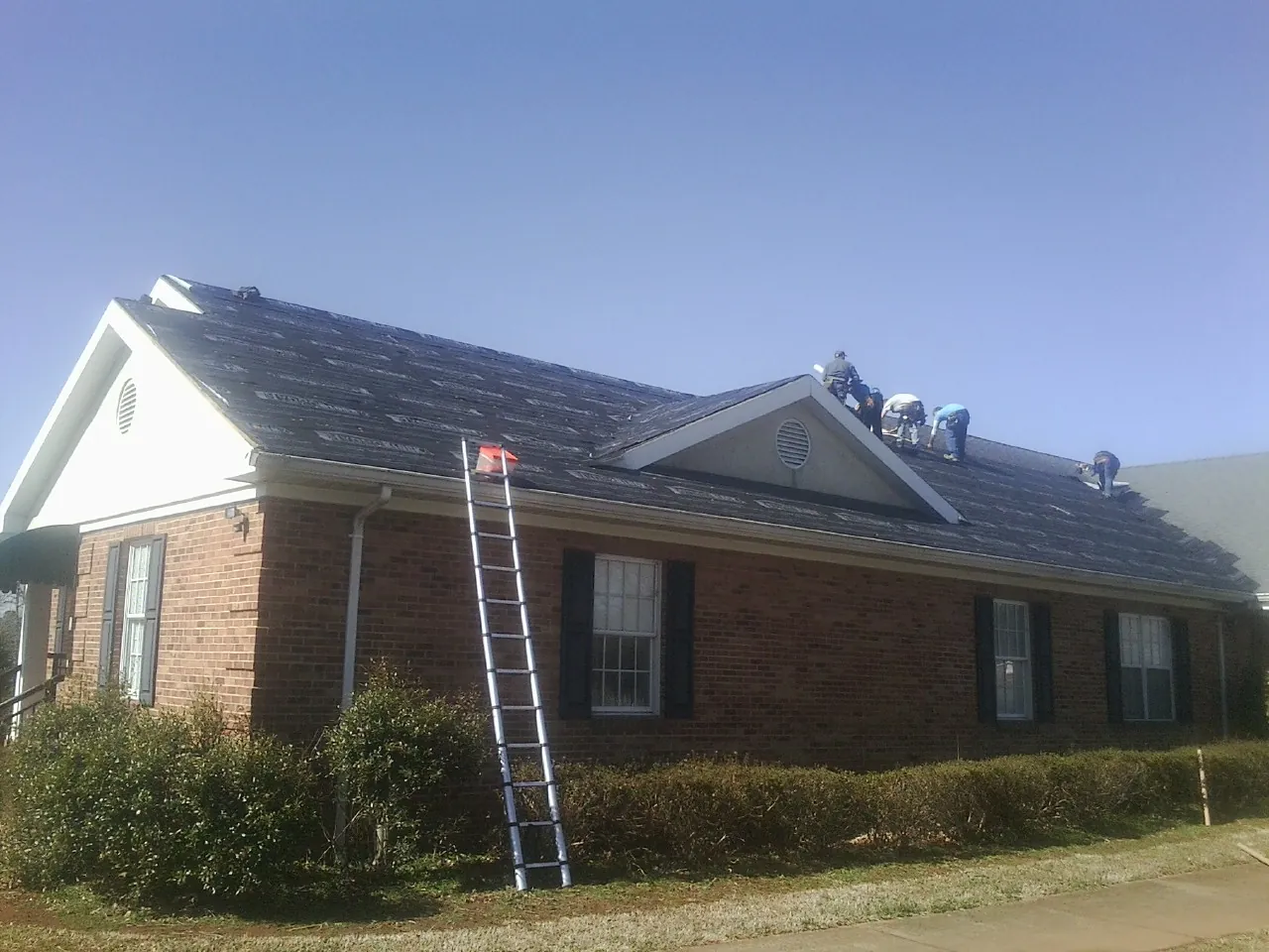 Crew tearing shingles off a brick ranch roof with a ladder set — Metal Roof Repair work by our crew in Chichester